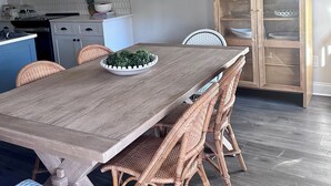 Zona pranzo con tavolo in legno, sedie in vimini, uno sgabello blu e un lampadario decorativo.