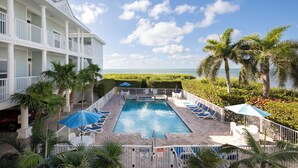 A pool area with lounge chairs and umbrellas, surrounded by palm trees and a view of the ocean.