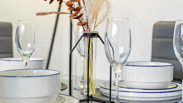 A dining table set with white plates and blue rimmed bowls, a clear glass vase with dried plants, and a tall glass candle holder.