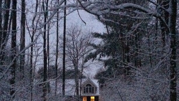 Un sentier enneigé menant à une maison dont une fenêtre est illuminée, le tout entouré d’arbres.