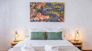 Een net opgemaakt bed met twee kussens, een stapel gevouwen handdoeken, en daarboven een schilderij van een kronkelende weg.