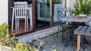 Eine Terrasse mit Tisch und Stühlen, im Hintergrund ein kleiner Küchenbereich durch Glastüren sichtbar, und Zimmerpflanzen in Töpfen.