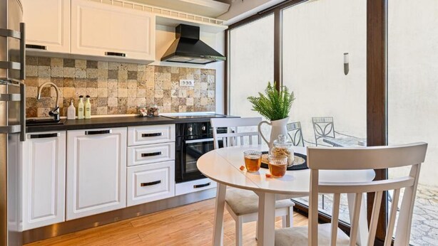 Eine moderne Küche mit weißen Schränken, einer schwarzen Dunstabzugshaube und einer gefliesten Spritzschutzwand. Es gibt eine Essecke mit einem weißen Tisch und Stühlen, und ein großes Fenster mit Blick.