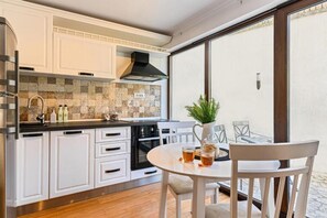 Cozinha moderna com armários brancos, coifa preta e backsplash em azulejo. Há uma área de jantar com mesa e cadeiras brancas, e uma grande janela com vista.