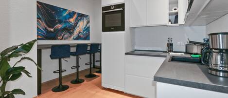A modern kitchen with white cabinets, a built-in oven, and a coffee maker. There are three blue bar stools and a large abstract painting on the wall.