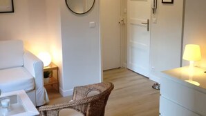A modern, well-lit living room with a white sofa, a wicker chair, a dining table, and a small side table with a lamp. - Charming 2 rooms old Lille, terrace, parking (Lille)