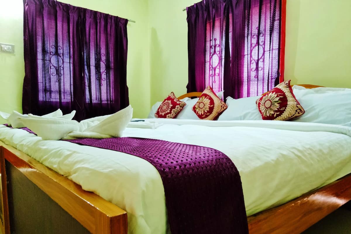 A neatly made bed with white linens and a purple blanket, red and gold patterned pillows, and purple curtains.