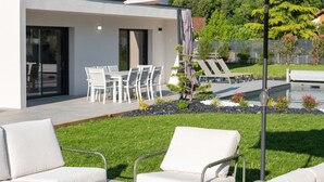 Un área de estar al aire libre moderna con mobiliario blanco, una piscina y montañas al fondo.