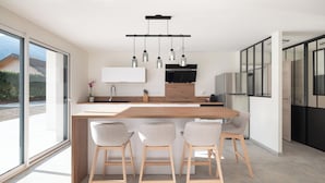 Una cocina moderna con una isla central, taburetes de bar, iluminación colgante y un ventanal grande.