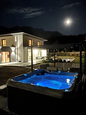 A hot tub with blue lighting, a covered patio area, and a house with illuminated windows. - Luxury Villa on Lake Annecy – Heated swimming pool & spa – Sleeps 8 (Doussard)