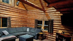L’intérieur d’une cabane en rondins, avec un canapé d’angle, un pouf et une petite table sur laquelle se trouve une lampe.