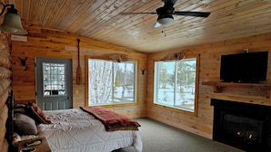 Une chambre dans un chalet en rondins, avec un lit, deux fenêtres, une cheminée et un ventilateur de plafond.