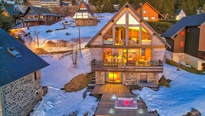 Una cabaña de madera de dos pisos con un frente de vidrio, un jacuzzi en el jardín delantero y un paisaje cubierto de nieve.