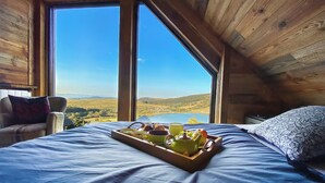 Un dormitorio con un ventanal grande, una cama, una bandeja con alimentos para el desayuno y vistas a un lago y colinas.