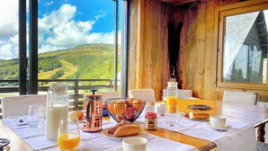 Una mesa de desayuno con vistas a colinas verdes, interior de madera y un ventanal amplio.