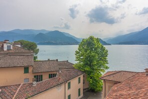 Vista de edificios con tejados de teja anaranjada y un árbol en primer plano, con vistas a un lago tranquilo y montañas al fondo.