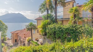 Un edificio sul lungomare con un balcone, immerso nel verde e con vista su un'acqua che si apre su montagne sullo sfondo.