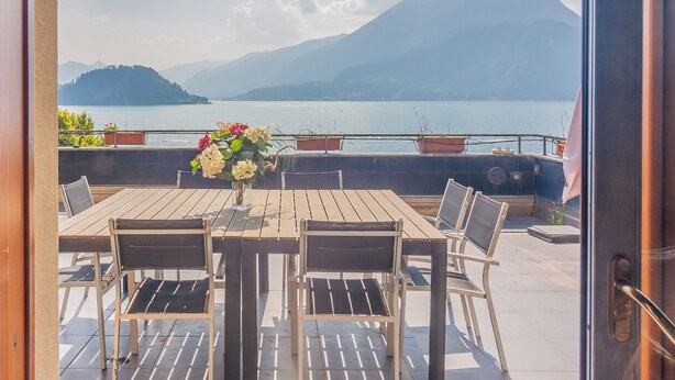 Un tavolo di legno con sedie e un vaso di fiori, con vista su un lago e montagne sullo sfondo.