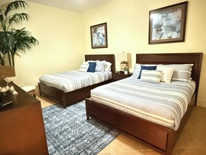 Une chambre d’hôtel avec deux lits simples, une tête de lit en bois, une table de chevet avec une lampe, une plante en pot et une petite table.