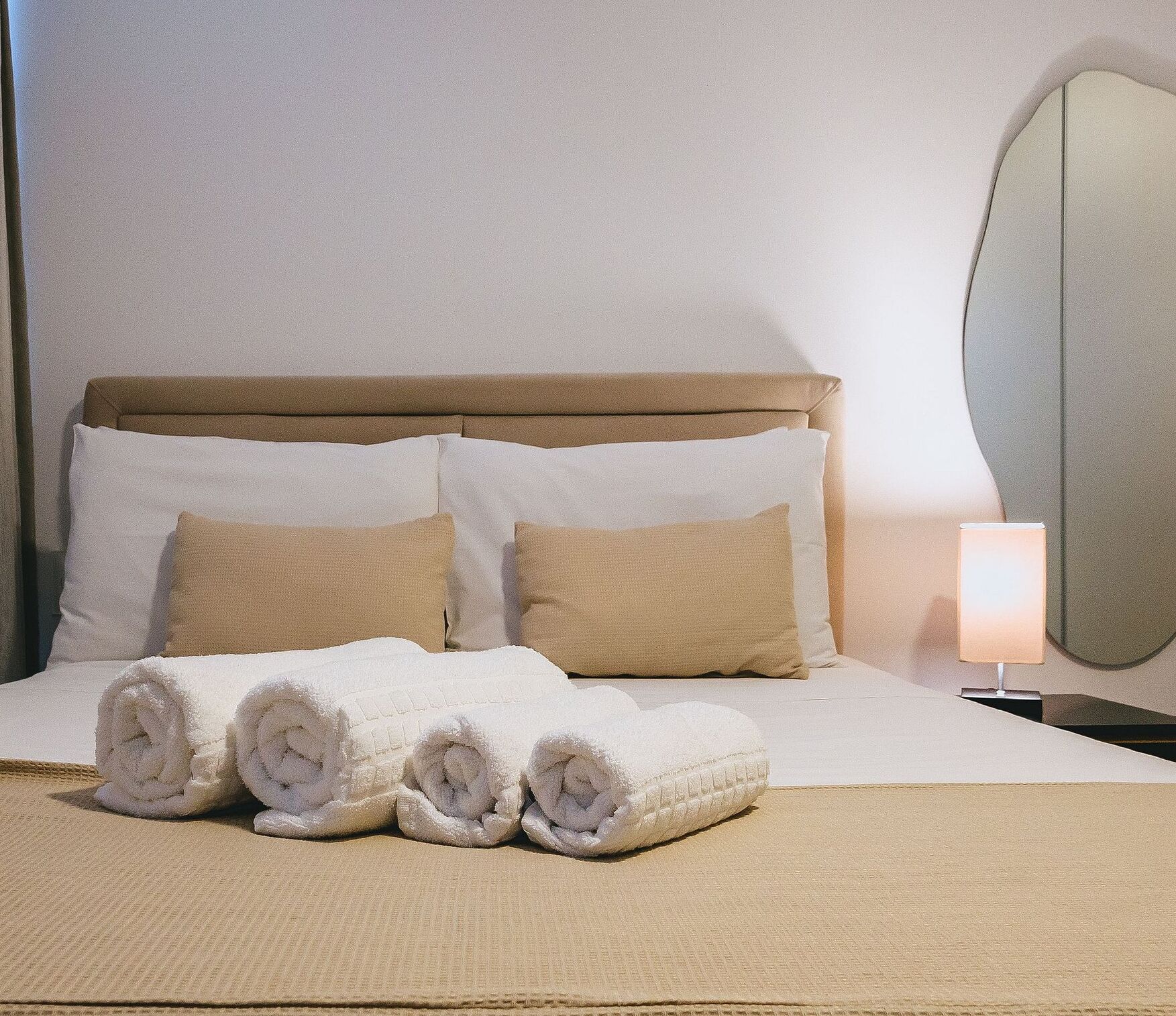 A neatly made bed with white and beige pillows, a rolled white towel, and a bedside lamp.