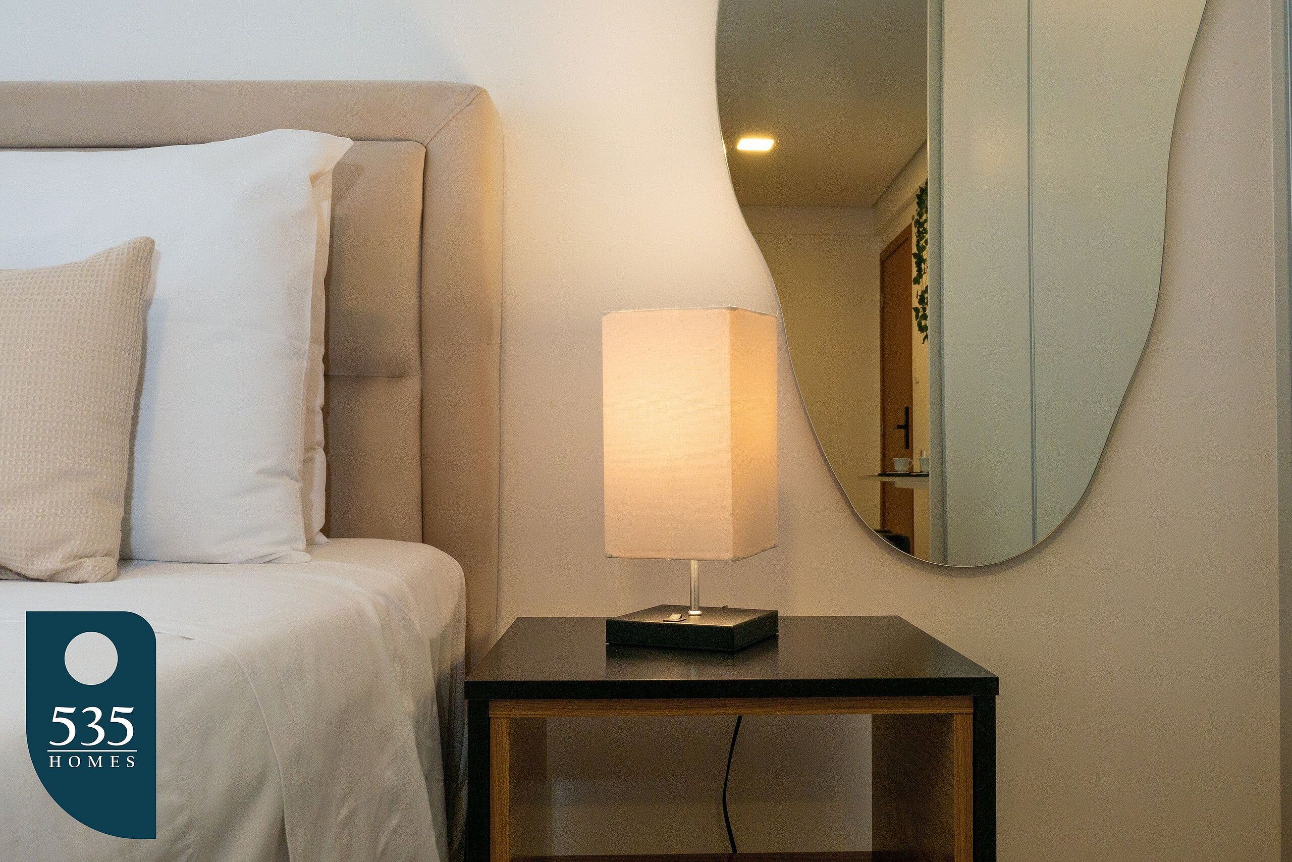 Ein ordentlich bezogenes Bett mit weißer Bettwäsche und einem beigen Kopfkissen, ein Nachttisch mit Lampe und ein runder Spiegel an der Wand.