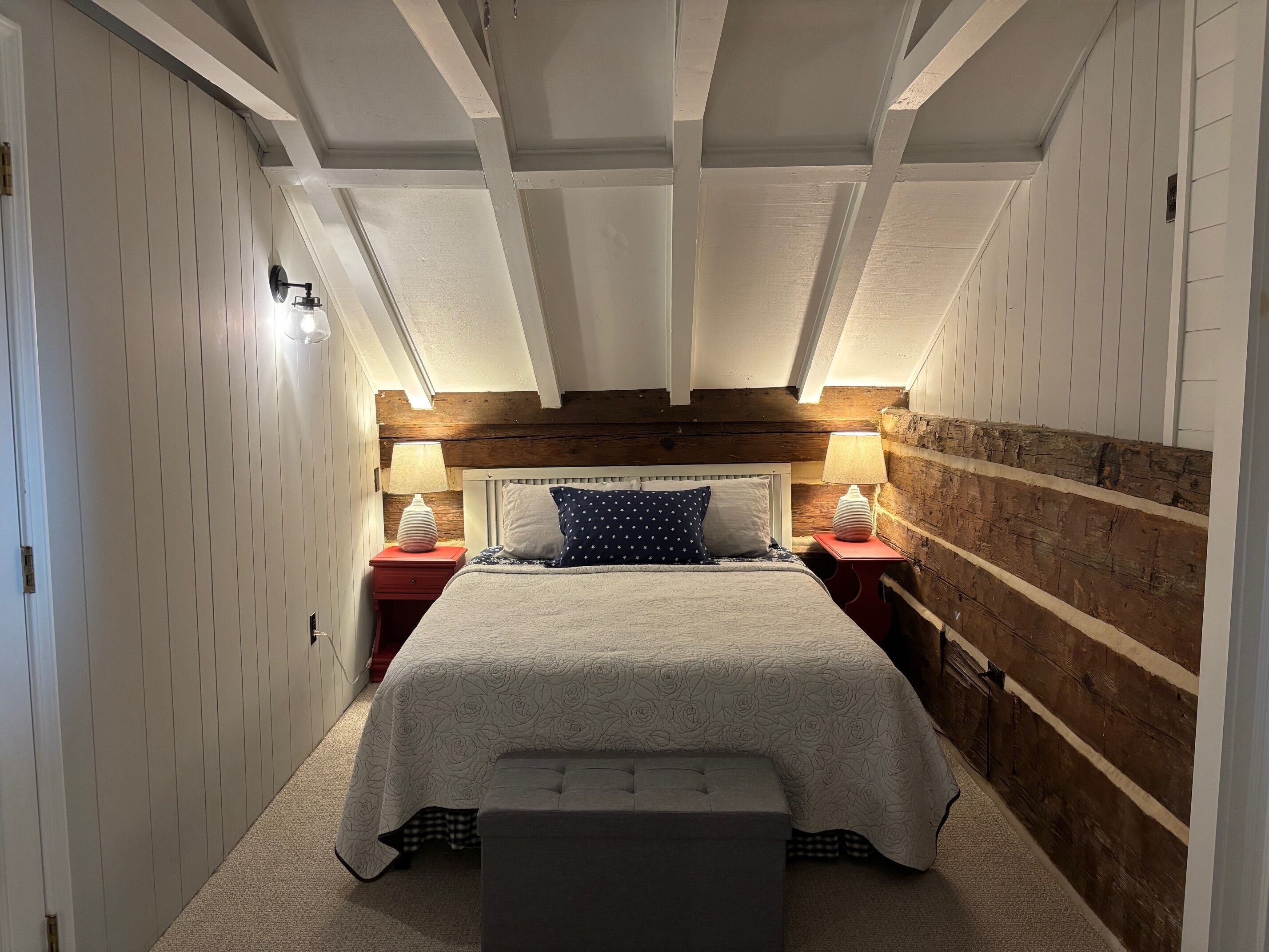 Um quarto com teto alto, vigas de madeira aparentes, uma cama grande com colcha florida, duas mesas de cabeceira com abajures e uma parede de destaque em madeira.