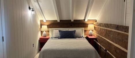 Um quarto com teto alto, vigas de madeira aparentes, uma cama grande com colcha florida, duas mesas de cabeceira com abajures e uma parede de destaque em madeira.