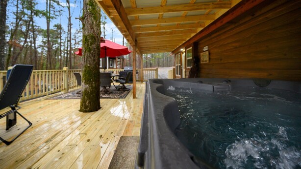 Una terraza de madera con hidromasaje, mobiliario exterior y vista a un bosque.
