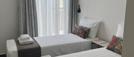 Ein Hotelzimmer mit zwei Betten, einem großen Fenster mit durchsichtigen Vorhängen, einem Nachttisch mit Lampe und einem an der Wand befestigten Telefon.