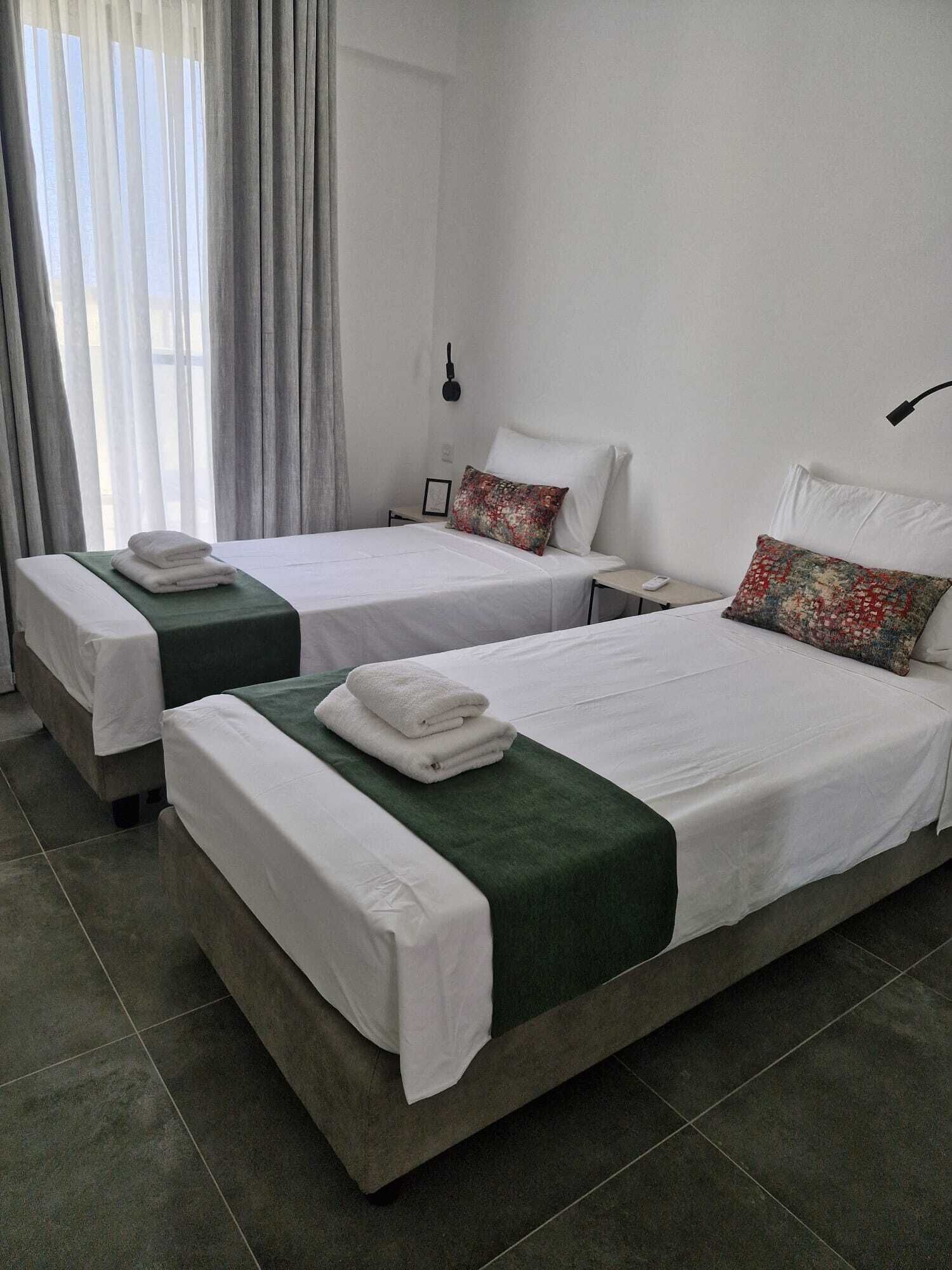 A hotel room with two beds, each with white linens and a green throw. There is a nightstand with a lamp, a window with curtains, and a view of the outside.