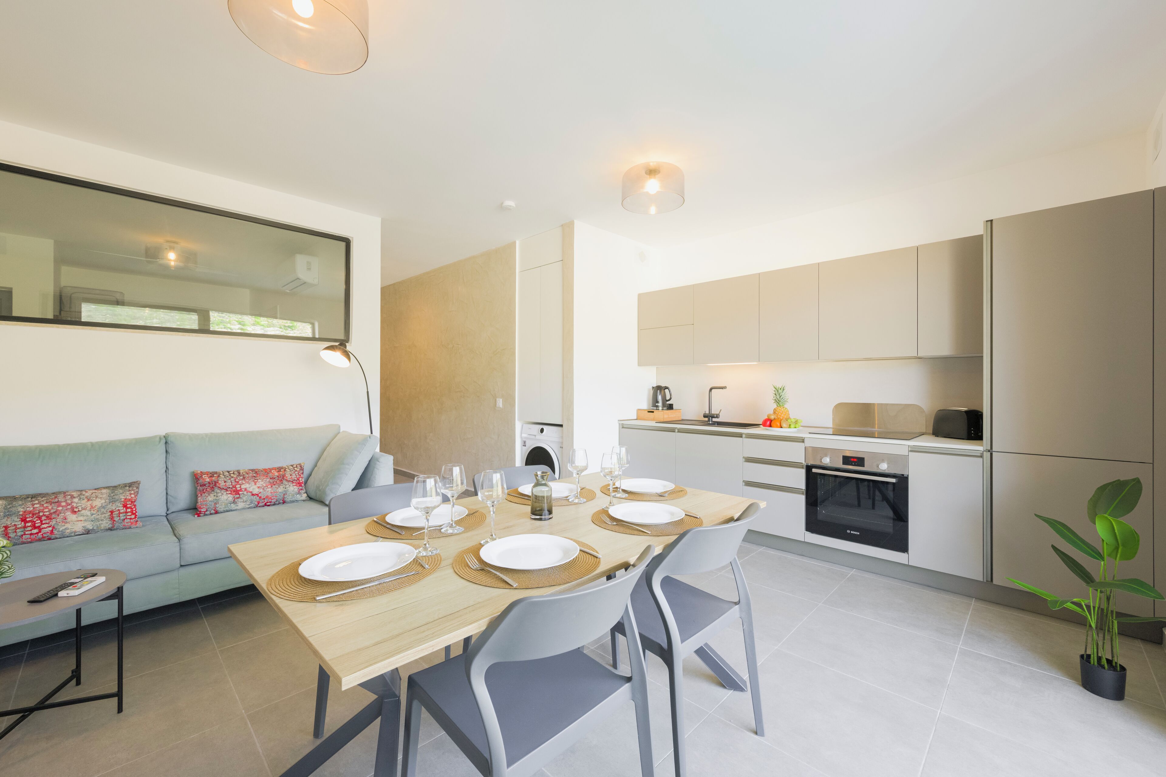 A modern kitchen with a dining area, a sofa, and a small table with remote controls.