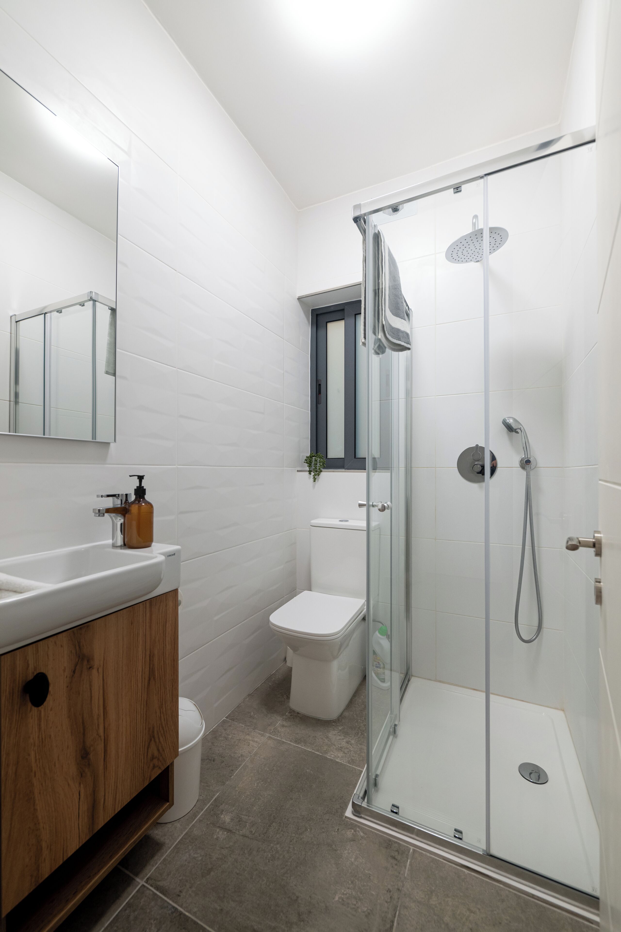 Et moderne badeværelse med en glasvæg omkring bruseren, et trækommode, et hvidt toilet og et spejl over vasken.