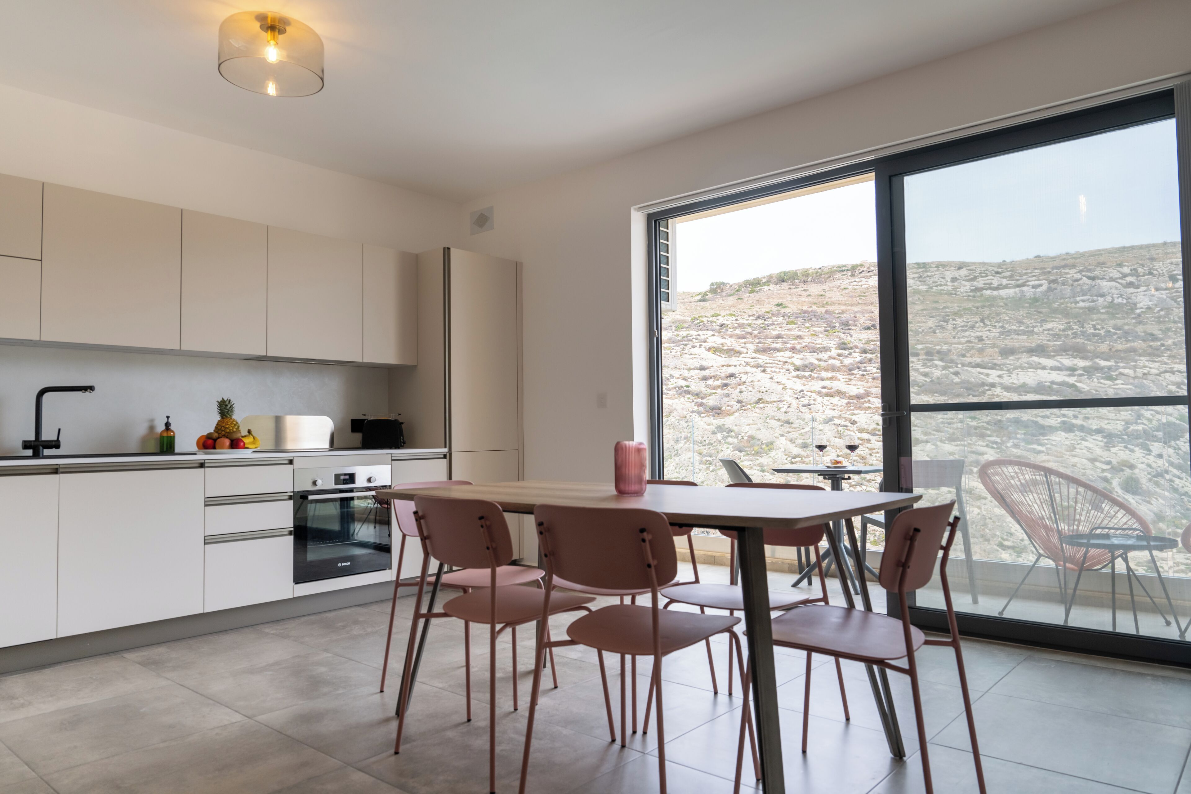 A modern kitchen with white cabinets, a large window, and a dining area with pink chairs.