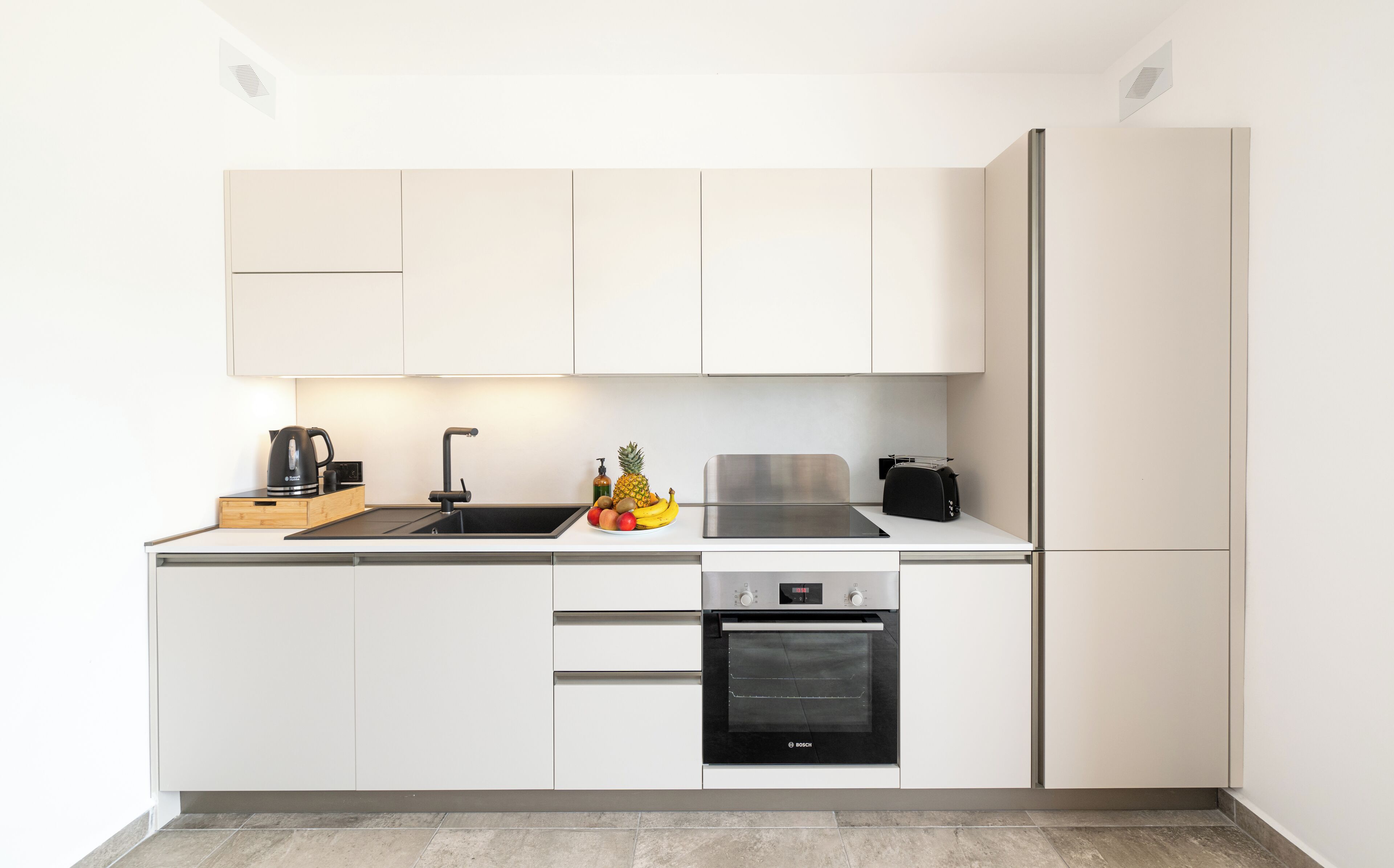 A modern kitchen with white cabinetry, a stainless steel sink, a Bosch oven, and a fruit bowl on the counter.