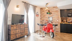 A modern living room with a TV, a wooden cabinet, a red scooter, and a kitchen area with dark cabinets and a brick wall. - VESPA SPRINT APARTMENT Amazing Nature 10' to the beach +PVT Park in City Centre! (Roverano Basso)