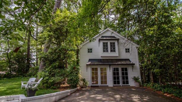 A white two-story house with a front porch, surrounded by lush greenery and trees. - Gorgeous Historic Carriage House with fine furnishings and modern amenities (Atlanta)