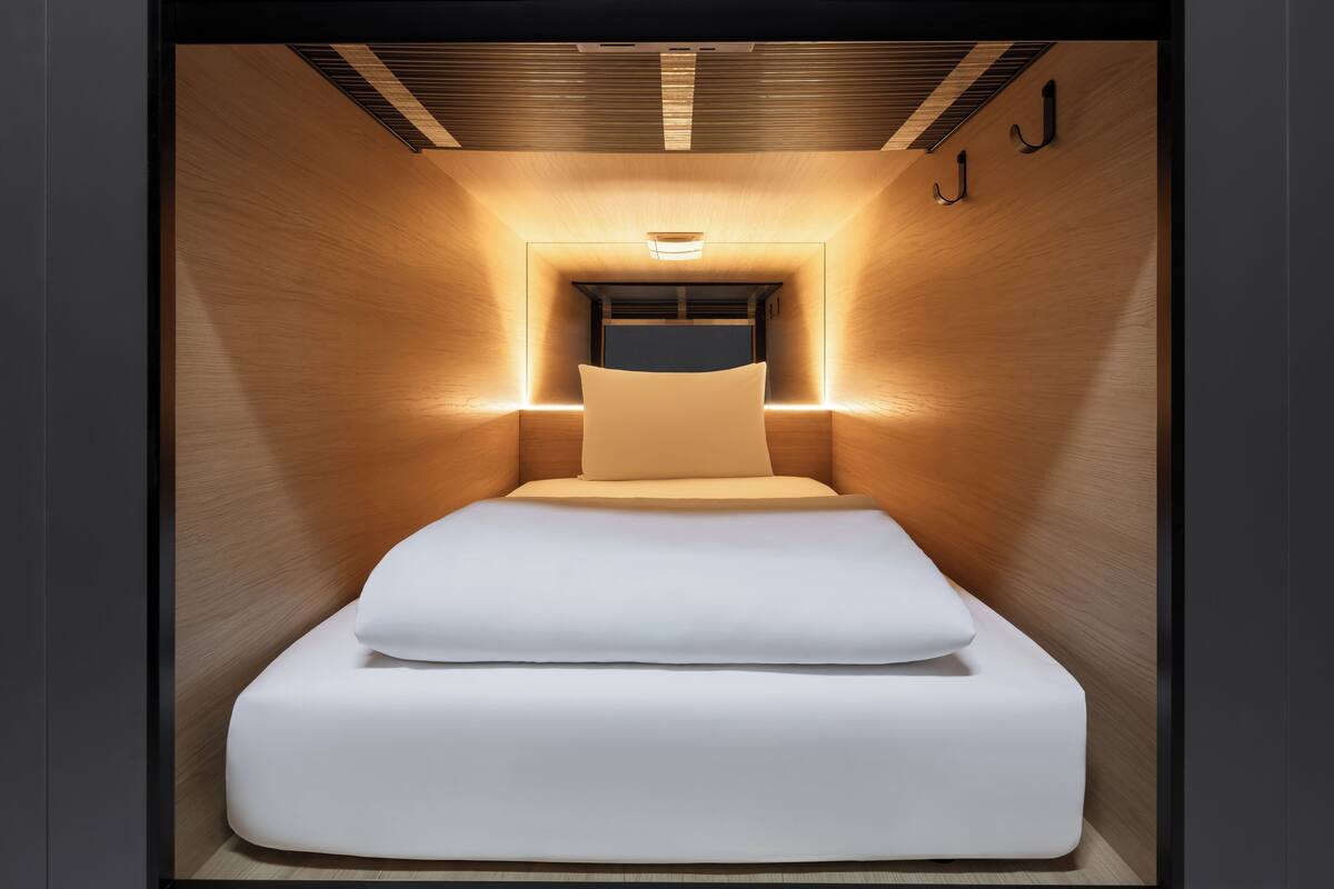 A modern, narrow hotel room with a bed centered in the middle, wooden walls, and a ceiling light fixture.