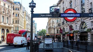 Una calle concurrida con una entrada de metro público, un autobús de dos pisos rojo y diversas tiendas.