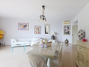 A living room with a white sofa, a dining table with chairs, a chandelier, and artwork on the walls.