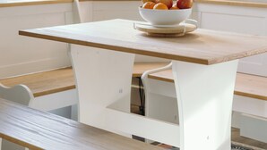 Ein Essbereich mit einem weißen Tisch und einer Bank, eine Obstschale auf dem Tisch und eine Lampe im Hintergrund.