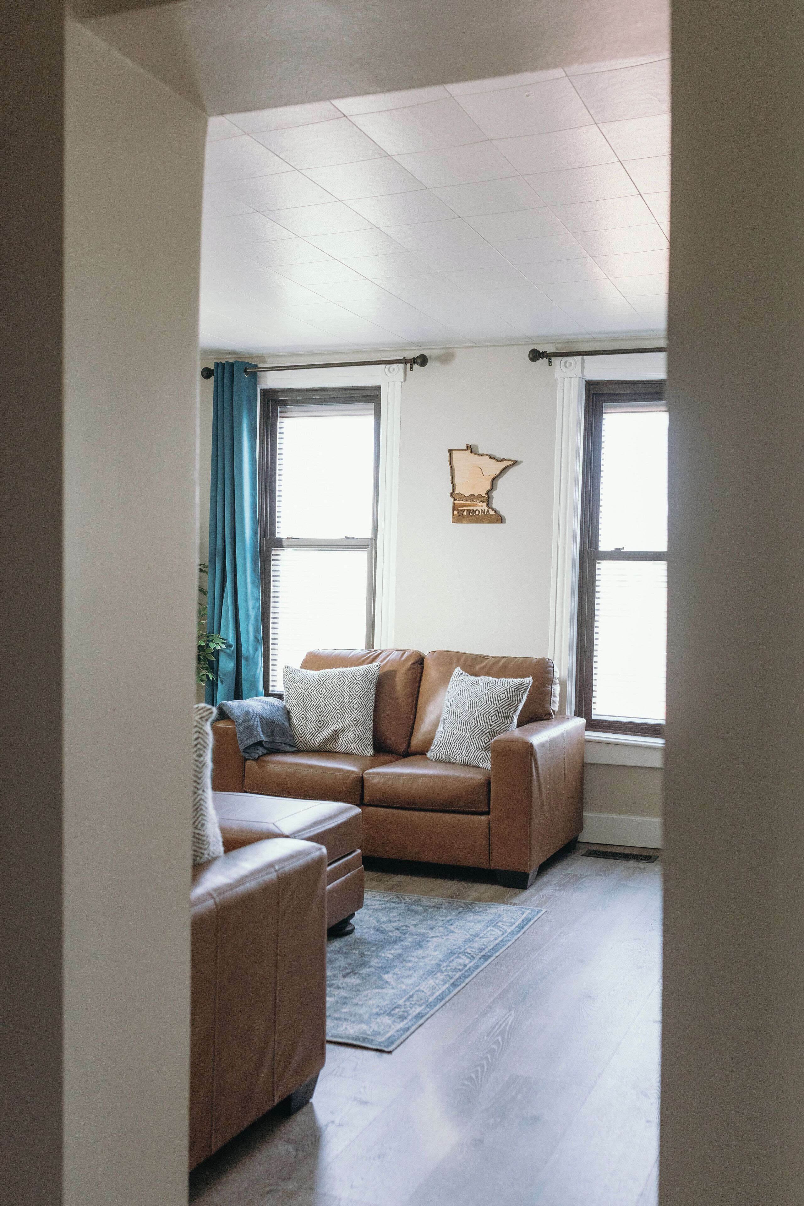 Ein Wohnzimmer mit einem braunen Ledersofa, einer gemusterten Tagesdecke und einem blauen Teppich. Zwei Fenster mit Vorhängen, eine Wandleuchte und eine gerahmte Karte an der Wand sind ebenfalls zu sehen.