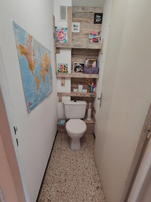 A small bathroom with a toilet, a wooden shelf with various items, a world map, and a sign that says "Home sweet home."