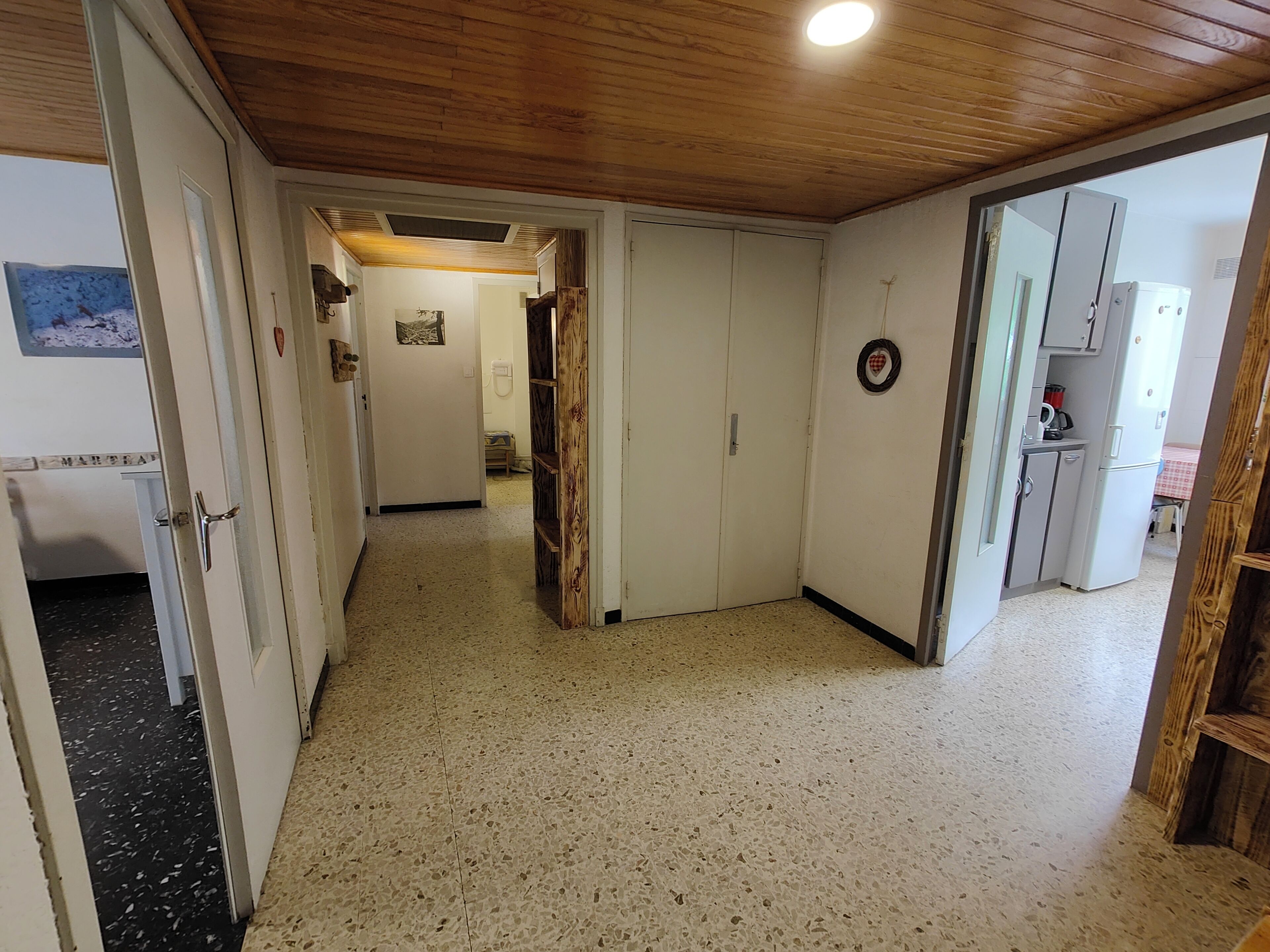 Un couloir avec un plafond en bois, un coin cuisine équipé d’un réfrigérateur, et une porte donnant sur une autre pièce.