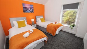 Et hotelværelse med to senge, begge med hvide sengetæpper og orange detaljer, et natbord med en lampe, et vindue med udsigt til grøn vegetation, og et maleri på den orange væg.
