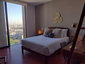 Een slaapkamer met een bed, nachtkastje, lamp en uitzicht op de stad door een groot raam.