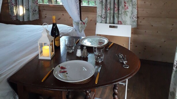 Una habitación de madera con una cama, una mesa dispuesta para comer y cortinas con motivos florales.