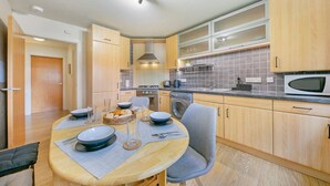 Een compacte keuken met houten kastjes, een magnetron, een fornuis en een spoelbak. Er staat een eettafel met stoelen en een schaal met fruit.