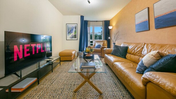 Een moderne woonkamer met een leren bank, een glazen salontafel, een flatscreen televisie en een groot raam met gordijnen.