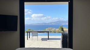 A bedroom with a large window offering a view of the sea and mountains. - Villa OLYMPE – 4 Bedrooms, Pool, Panoramic Sea View (Pietrosella)