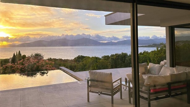 A modern outdoor lounge area with a glass balustrade overlooking a scenic view of a body of water and mountains during sunset. - Villa OLYMPE – 4 Bedrooms, Pool, Panoramic Sea View (Pietrosella)